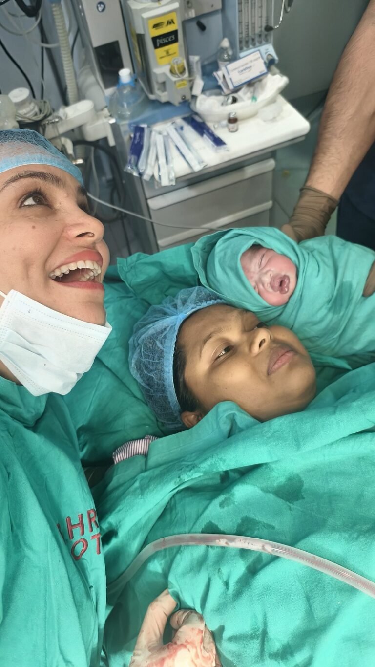 Dr. Archana Mohana in the delivery room with a joyful smile, a new mother, and a newborn baby moments after birth.
