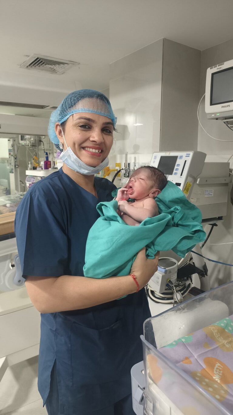 Dr. Archana Mohana smiling as she gently holds a newborn wrapped in a green cloth in a hospital neonatal care unit.