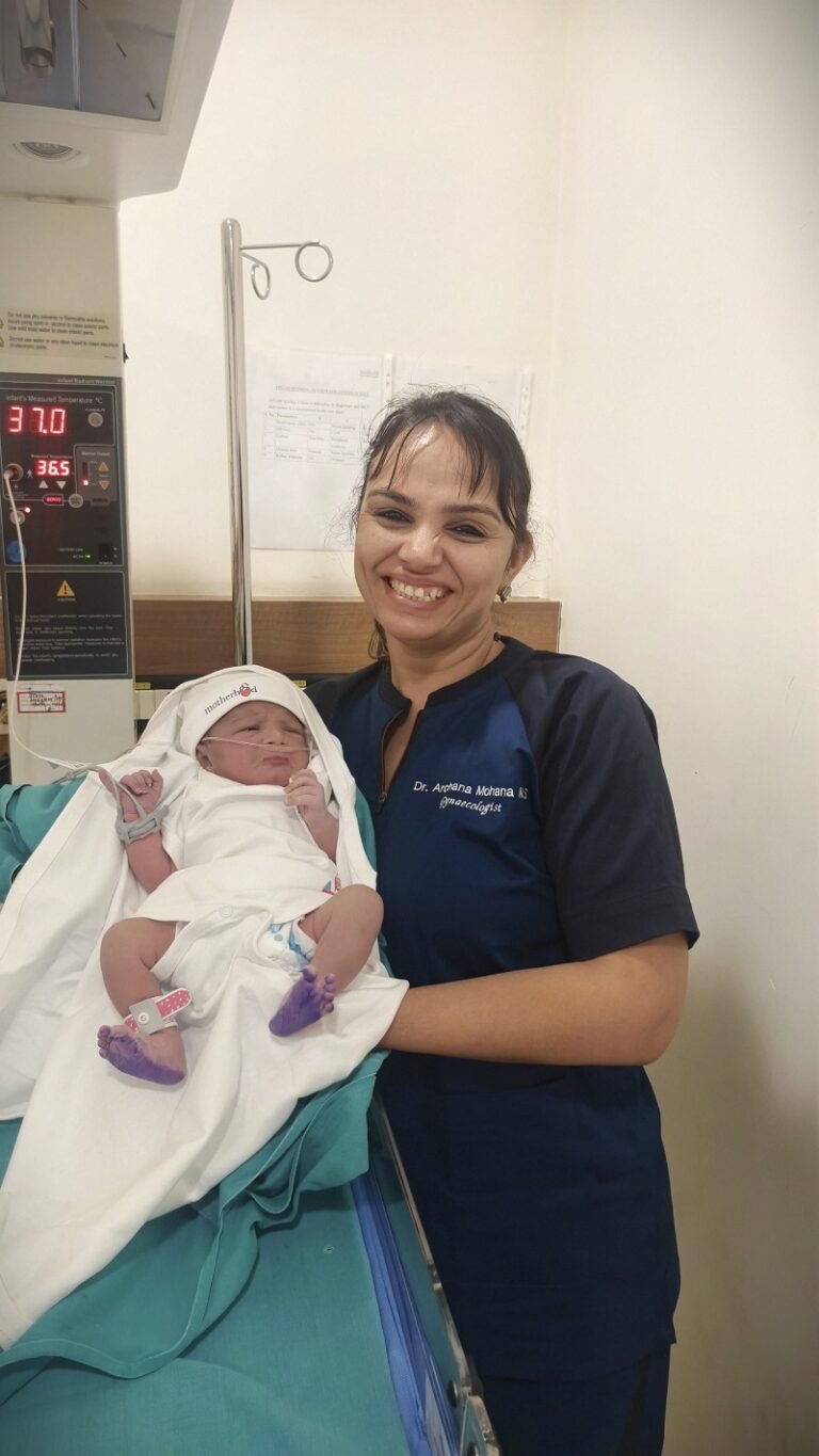 Dr. Archana Mohana, wearing a navy-blue medical uniform, smiles while gently holding a newborn wrapped in white at a hospital neonatal care unit.