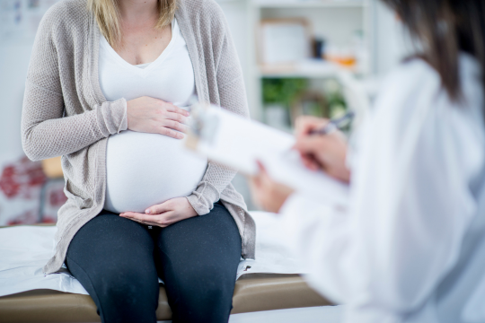 Pregnant woman sitting on a medical examination table, holding her belly, while a healthcare professional reviews notes on a clipboard.