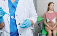 A gynecologist in a white coat and blue scrubs prepares a Pap smear test with a cervical brush and specimen container, while a female patient waits in the background on an examination chair.